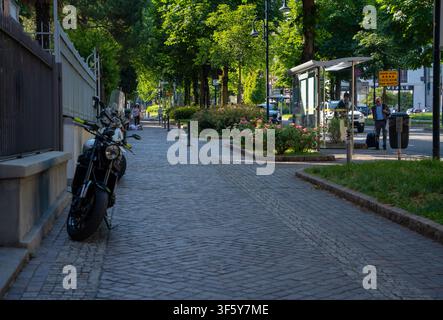 Bergamo, Italien – 16. Juni 2026: Helle Sommerstraßenszene in Bergamo mit einem geparkten Motorrad, gepflastertem Bürgersteig, üppigen grünen Bäumen und Glasbussh Stockfoto