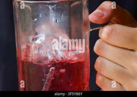 Ein gläserner Becher gefüllt mit rotem Eiskaltgetränk mit schmelzendem Eis und Hand, die den Griff hält, erfrischendes kaltes Getränk mit Kondenswasser und heller Farbe Stockfoto