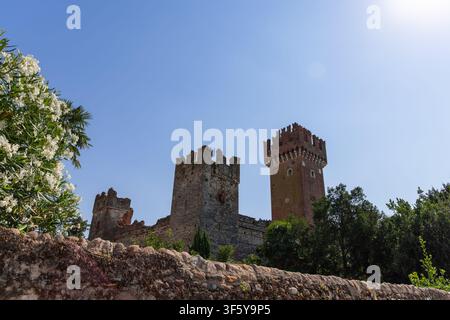 In Lazise, Italien, rahmen weiß blühende Sträucher und eine alte Grenz Mauer die Scaliger-Burg ein. Ihre zinnenbekrönten Backsteintürme heben sich deutlich von einem tiefen Blau ab Stockfoto