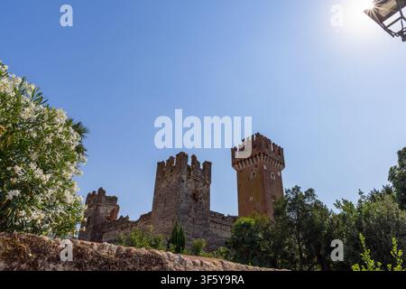 Helles Sonnenlicht umreißt die Ziegeltürme der Scaliger-Burg über den alten Mauern von Lazise, Italien. Grüne Bäume und ein klarer blauer Himmel umgeben das Mittelalterliche Stockfoto