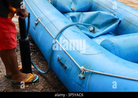 Ein aufblasbares blaues Floß, das am schlammigen Flussufer aufgeblasen wird, mit einer Person, die Luft pumpt, nasser Schlauchstruktur und befestigtem Seil, das Vorbereitung und Outdoor-Abenteuer vermittelt Stockfoto