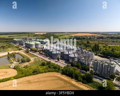 Hochwinkelaufnahme zeigt eine massive landwirtschaftliche Lageranlage mit Reihen von Stahlsilos neben grünen Feldern auf dem Land unter strahlender Sonne. Stockfoto