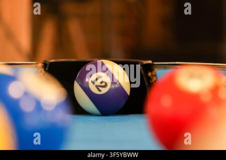 Ein lila und weiß gestreifter Billardball mit der Nummer zwölf, der in einer Tasche auf blauem Filz ruht, mit unscharfen bunten Bällen im Vordergrund, die eine verspielte Atmosphäre erzeugen Stockfoto
