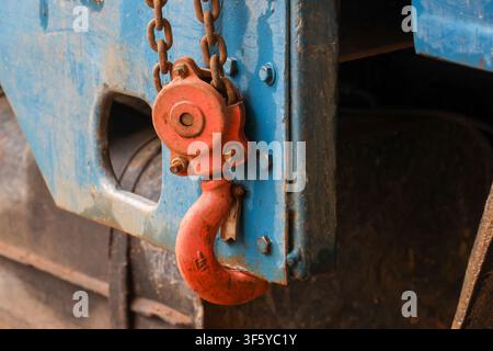 Ein rostroter Haken und eine Kette, die von blauer Metallmaschinerie hängen, Nahaufnahme eines industriellen Details, das Abnutzung, Textur, Bolzen und Farbkontrast zeigt, die Robustheit vermitteln Stockfoto