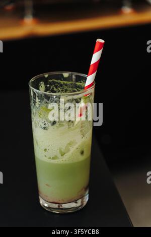 Nahaufnahme eines halb fertigen Eismatcha-Lattes mit einem gestreiften Papierstrohhalm. Erfrischendes grünes Teegetränk über Eis in einem modernen Café serviert. Stockfoto