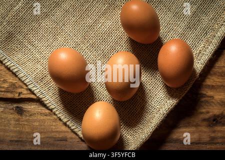 Fünf perfekte braune Eier auf einem Hintergrund aus Leinen und Holz; die Texturen werden in klaren Details dargestellt und durch den Einsatz von Licht und Schatten betont. Stockfoto