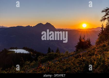 Ein atemberaubender Panoramablick auf einen Sonnenuntergang über den Alpen. Die leuchtend orangefarbene Sonne geht hinter einer Kette majestätischer, schneebedeckter Berge unter und wirft Stockfoto