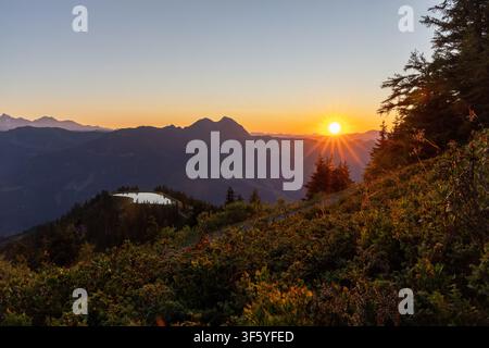 Ein atemberaubender Panoramablick auf einen Sonnenuntergang über den Alpen. Die leuchtend orangefarbene Sonne geht hinter einer Kette majestätischer, schneebedeckter Berge unter und wirft Stockfoto