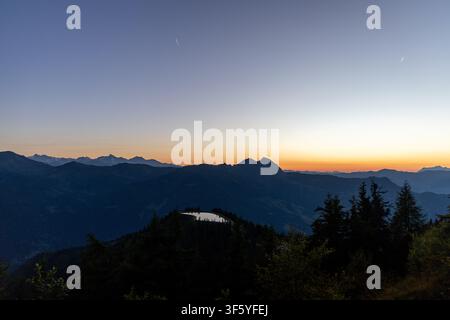 Ein atemberaubender Panoramablick auf einen Sonnenuntergang über den Alpen. Die leuchtend orangefarbene Sonne geht hinter einer Kette majestätischer, schneebedeckter Berge unter und wirft Stockfoto