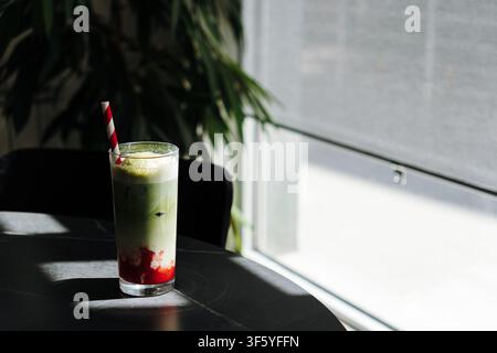 Schichtweiser Eistraum-Erdbeer-Matcha-Latte auf einem Cafétisch mit dramatischem Sonnenlicht von einem nahen Fenster. Erfrischendes handgefertigtes Getränk in einem modernen Stockfoto