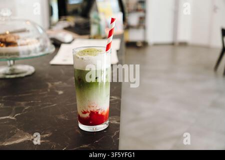 Schichtweiser Erdbeer-Matcha-Latte mit cremiger Sahnehaube, serviert mit einem gestreiften Papierstrohhalm. Erfrischendes Spezialkaffegetränk im Nahaufnahme. Stockfoto