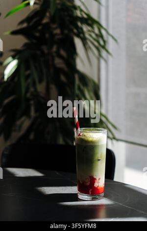 Schichtweiser Eistraum-Erdbeer-Matcha-Latte auf einem Cafétisch mit dramatischem Sonnenlicht von einem nahen Fenster. Erfrischendes handgefertigtes Getränk in einem modernen Stockfoto
