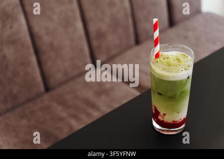Schichtweiser Erdbeer-Matcha-Latte serviert über Eis mit cremigem Schaum und einem gestreiften Papierstrohhalm. Erfrischendes Spezialgetränk, präsentiert in einem modernen Café Stockfoto