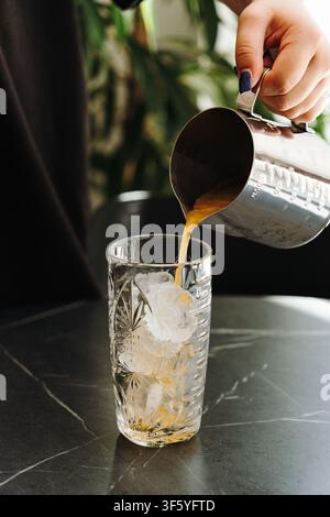 Frischer Orangensaft wird über Eiswürfel in ein dekoratives Glas gegossen. Erfrischendes Zitrusgetränk, zubereitet in einem modernen Café. Stockfoto