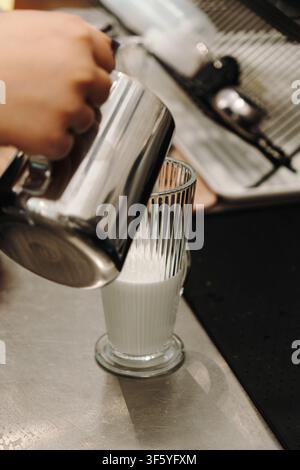 Frisch aufgeschäumte Milch wird während der Kaffeezubereitung in ein hohes Glas gegossen. Minimale Nahaufnahme, die einen professionellen Café-Arbeitsablauf einfängt. Stockfoto
