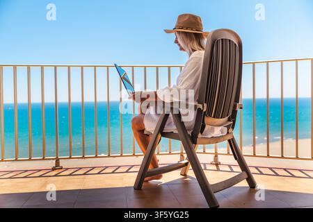 Frau nutzt Laptop, während sie sich während des Sommerurlaubs auf einem Balkon am Meer mit Meerblick und Fernarbeit entspannt Stockfoto