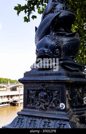 Dekorative Fischskulptur auf einem verzierten Laternenpfahl im viktorianischen Stil entlang der Themse in London, England, mit sichtbar dem Fluss und der Brücke Stockfoto