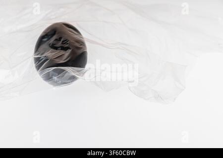 Nahaufnahme minimaler Studio-Stillleben eines einzelnen schwarz bemalten Eies, das in zerknitterter transparenter Plastikfolie auf einem weißen Hintergrund eingewickelt ist. Das einsame Ei umschließt Stockfoto