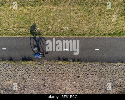 Luftaufnahme eines Radfahrers, der entlang einer asphaltierten Straße fährt, mit einem langen Schatten, der auf den Boden fällt, begrenzt von einem grasbewachsenen Straßenrand und einem stein gepflasterten Damm i Stockfoto