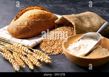 Handwerksbrot oder traditionelles Weizen-Rustikalbrot mit Mehl und Weizenähren auf dunklem Küchentisch, selektive Schärfentiefe. Stockfoto