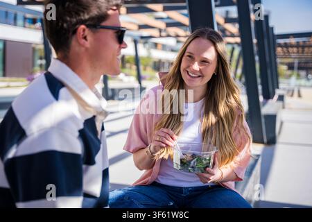 Paar lacht zusammen, während es einen gesunden Salat auf einer Bank unter einer Holzpergola an einem Tag isst Stockfoto