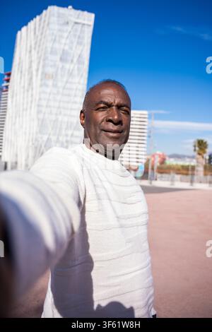 Reifer Mann, der ein lächelndes Selfie vor einem auffälligen modernen Gebäude an einem sonnigen Tag macht Stockfoto