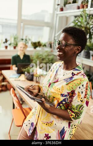 Vertikales Bild einer lächelnden jungen afroamerikanischen Frau, die ein Digitales Tablet in einem kreativen Büro hält, das vielfältige Zusammenarbeit am Arbeitsplatz und moderne Geschäftstechnologie repräsentiert Stockfoto