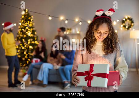 Glückliche Frau öffnet Weihnachtsgeschenkbox bei einem gemütlichen Heimparty mit Freunden und Familie Stockfoto