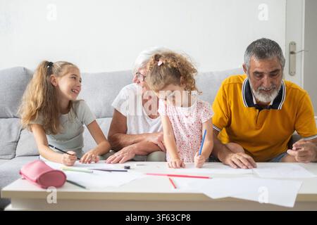 Zufriedene Großeltern, die Qualitätszeit mit ihren beiden Enkelinnen zu Hause verbringen und ihnen am Tisch beim Zeichnen und Lernen helfen. Stockfoto