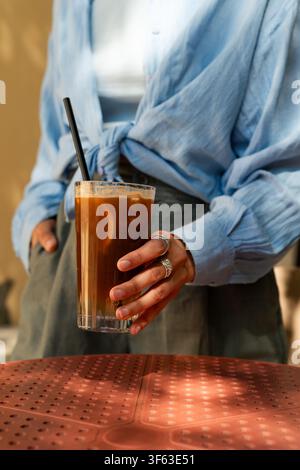 Nahaufnahme einer stilvollen Frau, die an einem Außentisch eines Cafés steht und ein gekühltes Espresso-Tonic-Getränk hält Stockfoto