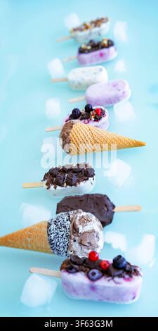 Bunte Runde aus hausgemachtem Joghurt- und Milch-Eis am Stiel. Vanille und Schokolade, verziert mit Nüssen und gefrorenen Beeren auf hellem blauem Hintergrund. Perfekt für eine sommerliche Kindergeburtstagsfeier. Stockfoto