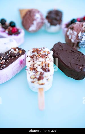 Bunte Runde aus hausgemachtem Joghurt- und Milch-Eis am Stiel. Vanille und Schokolade, verziert mit Nüssen und gefrorenen Beeren auf hellem blauem Hintergrund. Perfekt für eine sommerliche Kindergeburtstagsfeier. Stockfoto