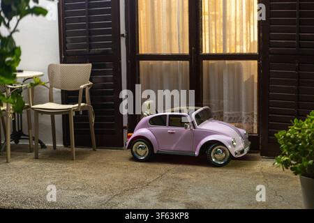 eine ruhige Seitenansicht eines lila Volkswagen Käfers, der in einer friedlichen Gartenumgebung geparkt ist, mit einem Stuhl und einem Tisch in der Nähe einer Metalltür und einer Holzkonstruktion Stockfoto