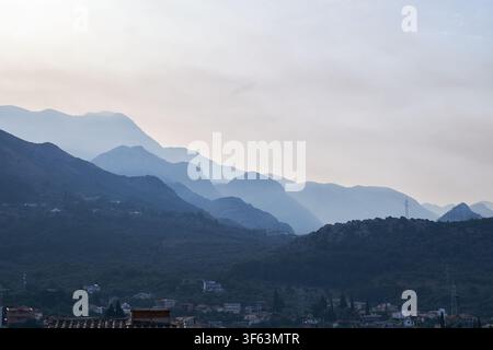 Schichtartige Gebirgszüge, die in Nebel über einer kleinen Stadt verschwinden und eine dramatische Landschaft unter einem bewölkten Himmel schaffen Stockfoto