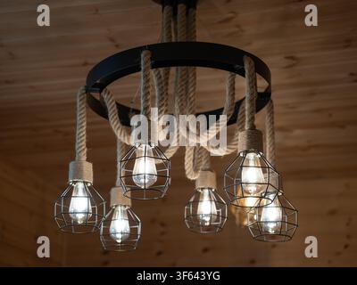 Rustikaler handgefertigter Kronleuchter beleuchtet eine Holzdecke, ausgestattet mit einem Metallring, Hanfschnüren und modernen Edison-Lampen, die in dekorativen Industrierahmen eingeschlossen sind Stockfoto