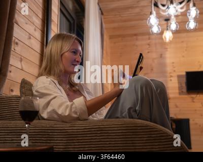 Frau entspannt sich auf einer Chaiselongue in einer gemütlichen Holzhütte, genießt die Freizeit, während sie ihr Smartphone durchstöbert und Rotwein trinkt Stockfoto