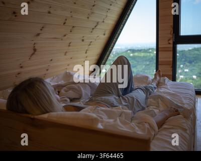Frau liegt bequem im Bett in einer gemütlichen A-Frame-Hütte und genießt den Panoramablick auf grüne Hügel, eine entfernte Stadt und das Meer, das von der L aus sichtbar ist Stockfoto