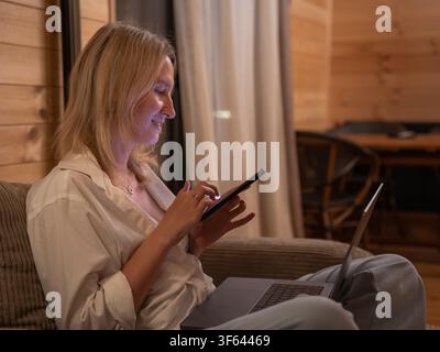 Blonde Frau lächelt, verwendet ein Smartphone, während sie auf einem bequemen Sofa sitzt, mit einem Laptop auf ihrem Schoß, und verwaltet persönliche Aufgaben oder Unterhaltung in einem c Stockfoto