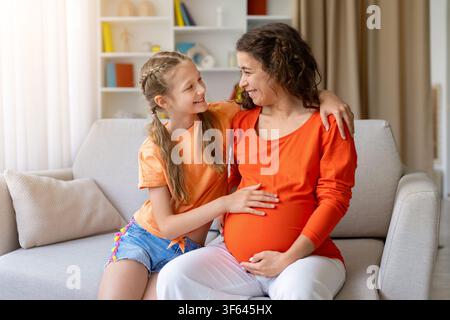 Lächelnde Tochter berührt sanft den Bauch der schwangeren Mutter, während sie zusammen auf dem Sofa sitzen Stockfoto