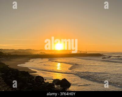 Atemberaubender Sonnenuntergang zur goldenen Stunde über einer felsigen Strandküste mit kleinen Booten in der Nähe des Wellenbrecher-Dammes in Pangandaran, Westjava, Indonesien. Stockfoto