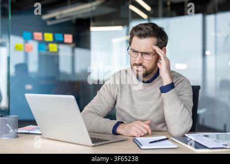Gestresster Geschäftsmann sitzt an einem Bürotisch, leidet unter Kopfschmerzen und Burnout-Symptomen, während er intensiv an einem Laptop arbeitet und frustriert ist Stockfoto