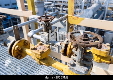 Nahaufnahme von verrosteten Handradventilen und gelben Rohren auf einem Stahlgittersteg in einer petrochemischen Industrieanlage, die gealterte Rohrverschraubungen und Va zeigt Stockfoto