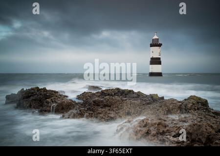 Der historische schwarz-weiß gestreifte Trwyn Du-Leuchtturm an der Penmon-Pointe, umgeben von einem seidigen, lang belichteten Meer und felsigem Vordergrund. Stürmisches Wetter Stockfoto