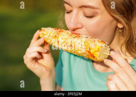 Nahaufnahme einer jungen Frau, die in gegrilltes mexikanisches Straßenmais elote mit Käse und Chili-Würze beißt, während sie einen sonnigen Imbiss im Freien genießt Stockfoto