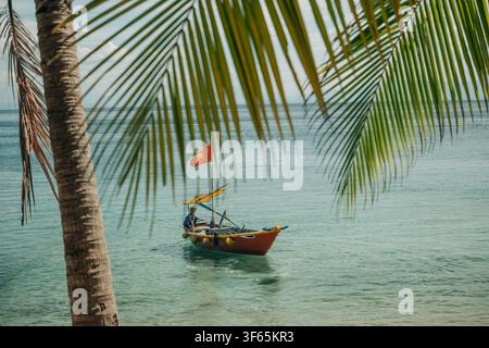 Hoi An, Vietnam, 15. Juli 2026. Ein motorisiertes Holzschiff treibt auf ruhigem türkisblauem Meerwasser, von der Küste aus durch das überhängende grüne Laub eingefangen Stockfoto