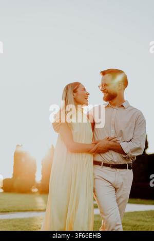 Ein Paar genießt einen Spaziergang und lacht gemeinsam bei Sonnenuntergang in einem Garten. Sie halten sich eng umschlungen und zeigen Glück, während die Sonne hinter ihnen untergeht. Stockfoto