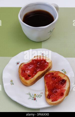 Vertikale Nahaufnahme von zwei süßen Toasts mit roter Himbeermarmelade auf einem Teller, serviert mit einer Tasse schwarzem Kaffee zum Frühstück zu Hause. Stockfoto