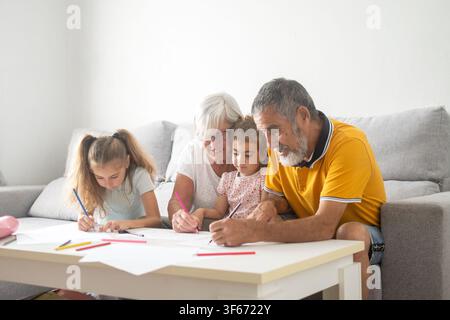 Zufriedene Großeltern, die Qualitätszeit mit ihren beiden Enkelinnen zu Hause verbringen und ihnen am Tisch beim Zeichnen und Lernen helfen. Stockfoto