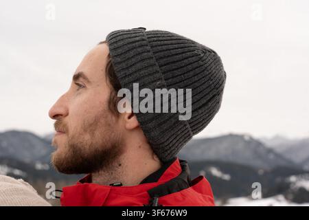 Seitenansicht Nahaufnahmeporträt eines nachdenklichen jungen bärtigen Mannes, der einen grauen Beanie-Mütze und eine rote Outdoor-Jacke trägt und in die Ferne blickt, auf einem Winterberg Stockfoto