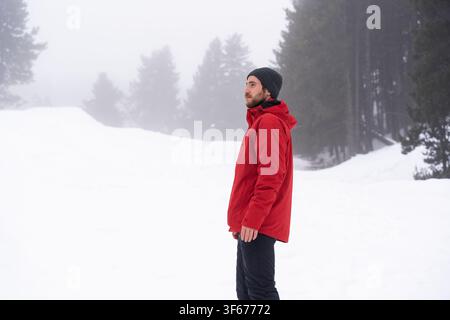 Junger Mann trägt eine rote Jacke und eine Mütze, steht in einer weiten schneebedeckten Landschaft mit Wald im Hintergrund und blickt ruhig in die neblige Ferne während Stockfoto